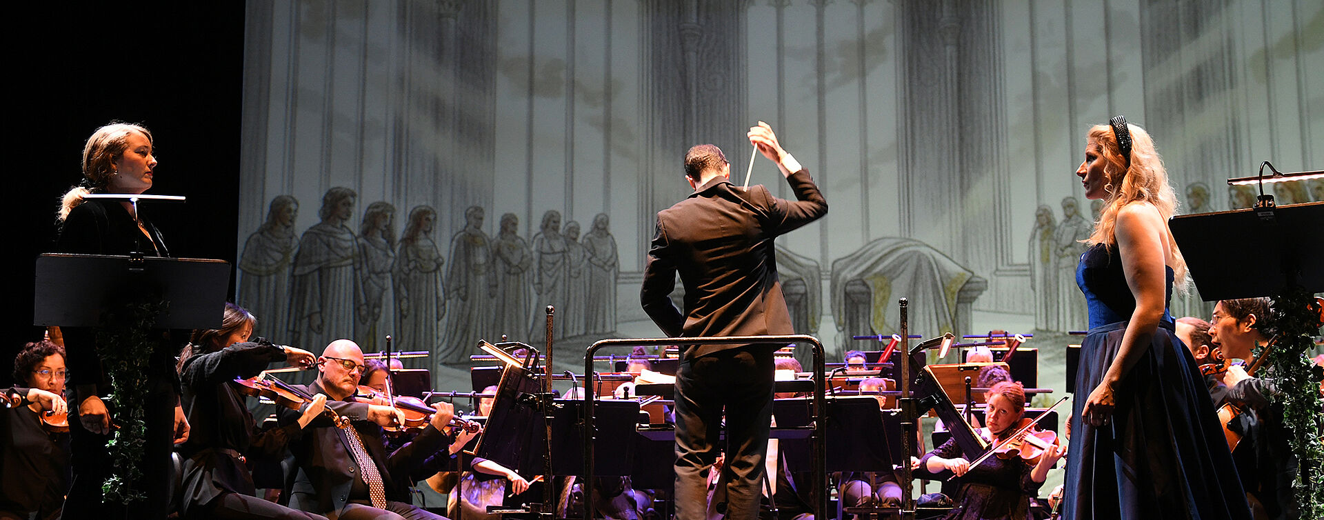 Im Vordergrund stehen zwei Personen, die sich ansehen. Dahinter ist das Orchester zu sehen. Als Hintergrundbild sind gezeichnete, graue Fenster erkennbar. 