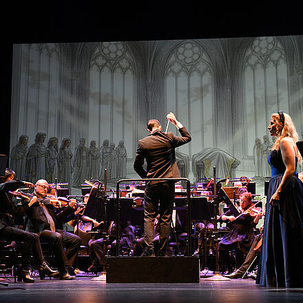 Im Vordergrund stehen zwei Personen, die sich ansehen. Dahinter ist das Orchester zu sehen. Als Hintergrundbild sind gezeichnete, graue Fenster erkennbar. 