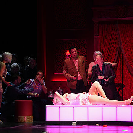 In der Mitte liegt eine Frau mit dem Rücken auf einem Podest. Um das Podest herum stehen mehrere Menschen.