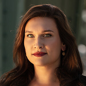 Portraitfoto von Boshana Milkov. Sie trägt einen roten Lippenstift und ihr braunes langes Haar offen. Dazu eine schwarte Bluse, darüber einen schwarzen Blazer. 