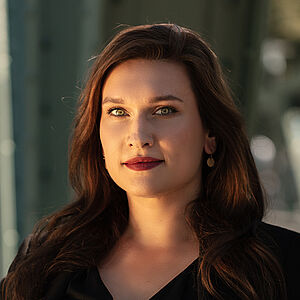 Portraitfoto von Boshana Milkov. Sie trägt einen roten Lippenstift und ihr braunes langes Haar offen. Dazu eine schwarte Bluse, darüber einen schwarzen Blazer. 