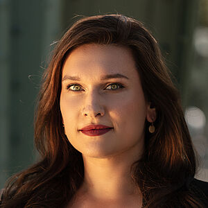 Portraitfoto von Boshana Milkov. Sie trägt einen roten Lippenstift und ihr braunes langes Haar offen. Dazu eine schwarte Bluse, darüber einen schwarzen Blazer. 