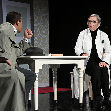 Im Vordergrund steht ein Tisch. An diesem Tisch sitzen ein Mann und eine Frau. Im Hintergrund steht ein zweiter Tisch, an dem eine Frau sitzt.