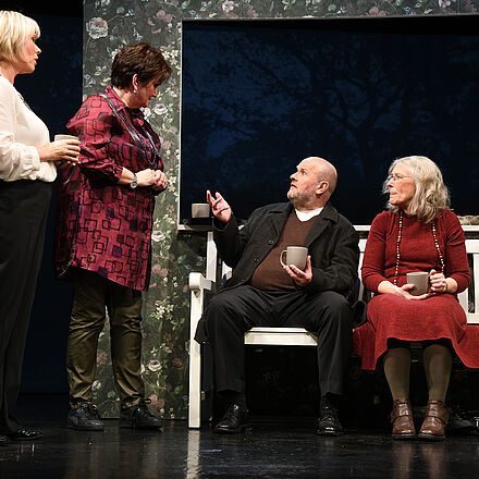 Ein Mann und eine Frau sitzen auf einer Bank, neben ihnen stehen zwei Frauen, sie sprechen zusammen.