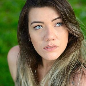 Portraitfoto von Lilian Giovanini. Sie schaut nach links oben. Ihre hellblauen Augen stechen hervor. Ihr langes braunes Haar mit blonden Strähnen fällt ihr über die Schultern. Der Hintergrund ist grün und verschwommen.