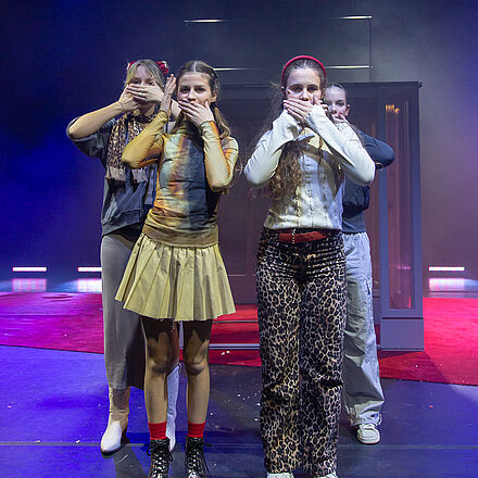 Alle halten sich den Mund zu und stehen vor einem viereckigen Gebäude.