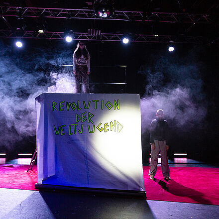 Das gesamte Ensemble steht auf der Bühne. Es weht eine Fahne „Revolution der Weltjugend“ 