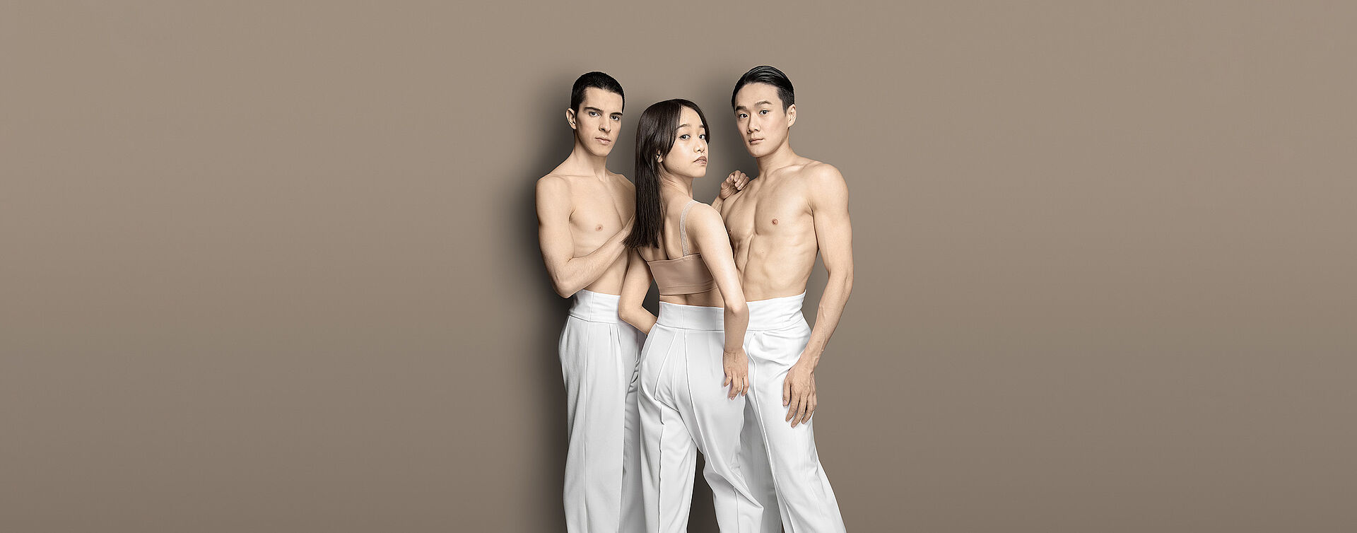 Eine Frau steht zwischen zwei Männern. Die Männer tragen nur eine weiße lange Hose und sind oberkörperfrei. Die beiden haben kurze dunkle Haare. Die Frau hat lange dunkle Haare und sie trägt ein hautfarbendes Bandeau und eine weiße lange Hose. Sie steht zu den Männern zugewandt und schaut über ihre Schulter den Betrachtenden an. Der Hintergrund ist goldfarbend.