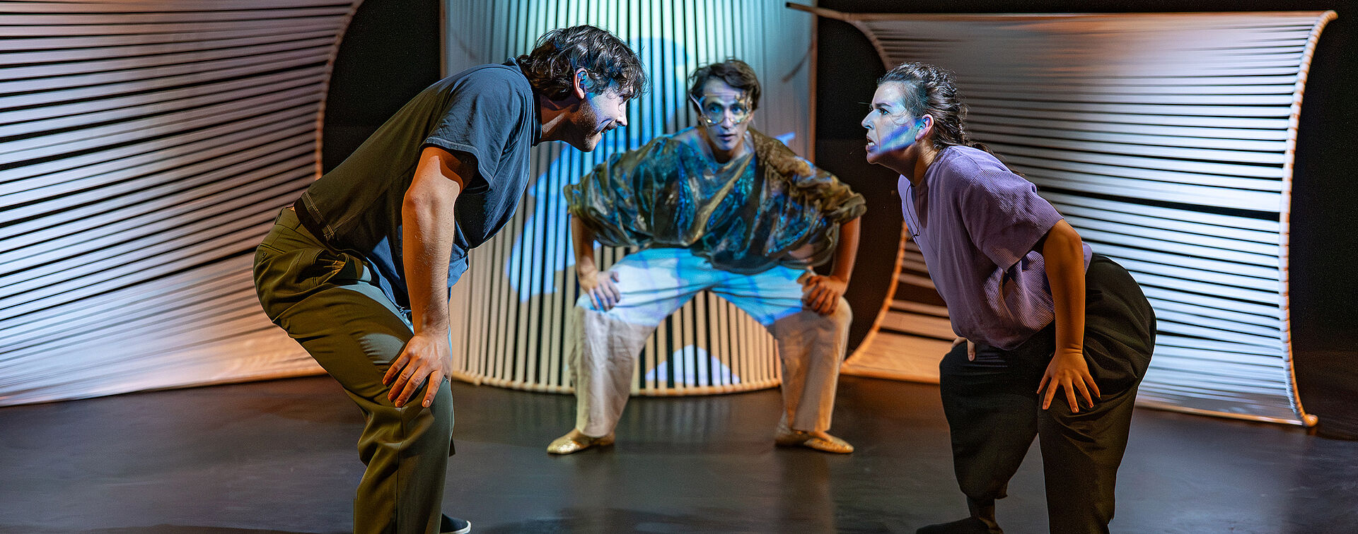 Drei Personen die leicht gebeugt stehen, sich auf den Knien abstützen und sich anschauen. Links und in der Mitte stehen Männer. Rechts steht die Frau. 