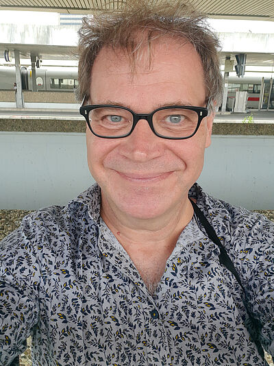 Portraitfoto von Patrick Schimanski. Er strahlt in die Kamera. Er trägt eine dunkle Brille und ein geblümtest Hemd.
