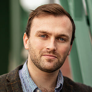 Portraitfoto von Andrew Irwin. Er trägt eine dunkelbraune karierte Anzugjacke, darunter ein blau-weiß kariertes Hemd. Die oberen Knöpfe sind geöffnet. Er umfasst mit dem linken Arm die Strebe eines grünen Brückenkonstrukts.