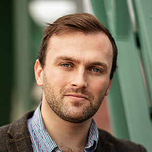 Portraitfoto von Andrew Irwin. Er trägt eine dunkelbraune karierte Anzugjacke, darunter ein blau-weiß kariertes Hemd. Die oberen Knöpfe sind geöffnet. Er umfasst mit dem linken Arm die Strebe eines grünen Brückenkonstrukts.