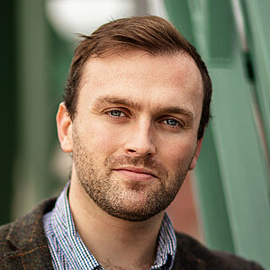 Portraitfoto von Andrew Irwin. Er trägt eine dunkelbraune karierte Anzugjacke, darunter ein blau-weiß kariertes Hemd. Die oberen Knöpfe sind geöffnet. Er umfasst mit dem linken Arm die Strebe eines grünen Brückenkonstrukts.