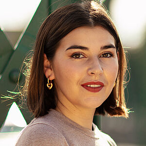 Portraitfoto von Coco Plümer. Sie steht vor grünen Stahlstreben. Sie steht seitlich und blickt nach vorn. Ihr braunes Haar ist schulterlang. Sie trägt goldene Creolen und roten Lippenstift. Ihr Oberteil ist beige.