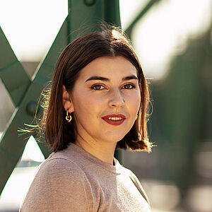 Portraitfoto von Coco Plümer. Sie steht vor grünen Stahlstreben. Sie steht seitlich und blickt nach vorn. Ihr braunes Haar ist schulterlang. Sie trägt goldene Creolen und roten Lippenstift. Ihr Oberteil ist beige.