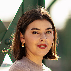 Portraitfoto von Coco Plümer. Sie steht vor grünen Stahlstreben. Sie steht seitlich und blickt nach vorn. Ihr braunes Haar ist schulterlang. Sie trägt goldene Creolen und roten Lippenstift. Ihr Oberteil ist beige.