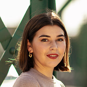 Portraitfoto von Coco Plümer. Sie steht vor grünen Stahlstreben. Sie steht seitlich und blickt nach vorn. Ihr braunes Haar ist schulterlang. Sie trägt goldene Creolen und roten Lippenstift. Ihr Oberteil ist beige.