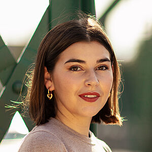 Portraitfoto von Coco Plümer. Sie steht vor grünen Stahlstreben. Sie steht seitlich und blickt nach vorn. Ihr braunes Haar ist schulterlang. Sie trägt goldene Creolen und roten Lippenstift. Ihr Oberteil ist beige.