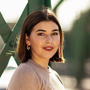 Portraitfoto von Coco Plümer. Sie steht vor grünen Stahlstreben. Sie steht seitlich und blickt nach vorn. Ihr braunes Haar ist schulterlang. Sie trägt goldene Creolen und roten Lippenstift. Ihr Oberteil ist beige.
