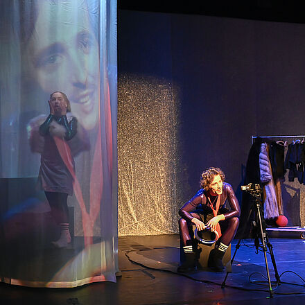 Hinter dem Vorhang posiert eine Person. Neben ihr sitzt eine Person auf dem Hocker und schaut in die Kamera. Das Bild wird auf den Vorhang projiziert. Im Hintergrund erkennt man Kleidung.