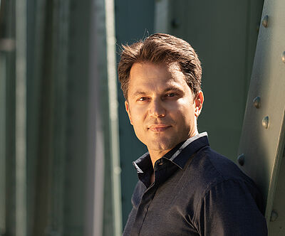Portraitfoto von Konstantinos Klironomos. Er trägt ein dunkelblaues Hemd und steht seitlich an eine grüne Stahlstrebe gelehnt.