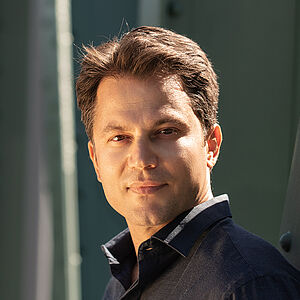 Portraitfoto von Konstantinos Klironomos. Er trägt ein dunkelblaues Hemd und steht seitlich an eine grüne Stahlstrebe gelehnt.
