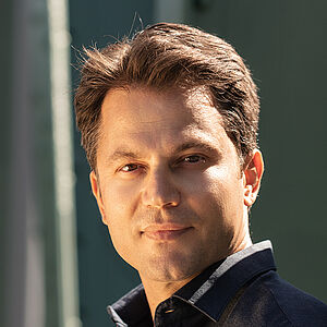 Portraitfoto von Konstantinos Klironomos. Er trägt ein dunkelblaues Hemd und steht seitlich an eine grüne Stahlstrebe gelehnt.