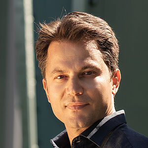 Portraitfoto von Konstantinos Klironomos. Er trägt ein dunkelblaues Hemd und steht seitlich an eine grüne Stahlstrebe gelehnt.