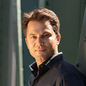 Portraitfoto von Konstantinos Klironomos. Er trägt ein dunkelblaues Hemd und steht seitlich an eine grüne Metallstrebe gelehnt.