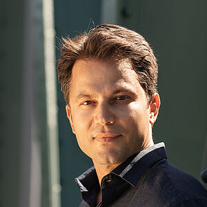 Portraitfoto von Konstantinos Klironomos. Er trägt ein dunkelblaues Hemd und steht seitlich an eine grüne Stahlstrebe gelehnt.