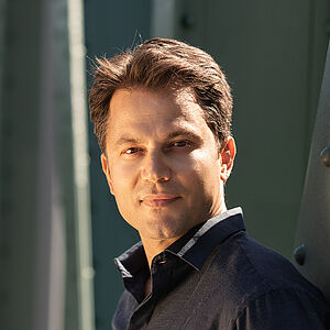 Portraitfoto von Konstantinos Klironomos. Er trägt ein dunkelblaues Hemd und steht seitlich an eine grüne Stahlstrebe gelehnt.