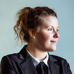 Portraitfoto von Annalena Hösel mit verschränkten Armen. Sie blickt zur Seite. Sie ist in einen Anzug gekleidet. Darunter trägt sie ein weißes Hemd und ein schwarzes gebundenes Tuch. Ihr lockiges Haar ist locker hochgesteckt. 