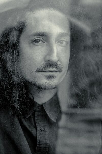 Schwarzweißes Portraitfoto von Nikolaj Efendi. Er hat langes welliges Haar und trägt einen Schurrbart sowie einen dezenten Kinnbart. Er blickt durch eine Glasscheibe. Er ist in ein zugeknöpftes grobes Hemd.