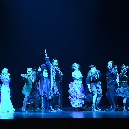 Zu sehen ist eine Gruppe von Menschen, die alle eine Hand nach vorne strecken. Der Protagonist steht im Mittelpunkt des Bildes und hält eine Rasierklinge in der Hand.