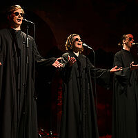 Drei Sänger:innen in schwarzen Kutten mit schwarzen Sonnenbrillen. Sie halten ihre Arme vor sich ausgestreckt und singen in Mikrofone. Die Bühne ist dunkel.