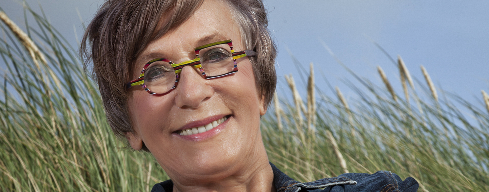 Portraitfoto von Gisa Pauly. Sie grinst in die Kamera. Ihr kurzes Haar wehnt im Wind. Sie trägt eine bunte Brille, dessen eines Glas rund und das andere quadratisch ist. Im Hintergrund weht Schilfrohr. Der Himmel ist blau-grau.