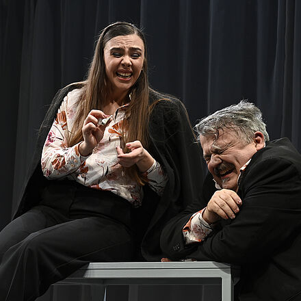 Eine Frau sitzt auf einem Tisch und hält einen aufgerollten Geldschein in der Hand. Ein Mann sitzt neben dem Tisch und stützt sich darauf ab. Beide Personen lachen.