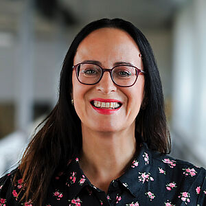 Portraitfoto von Iris Wemme-Baranowski. Sie lächelt mit offenem Mund. Ihr dunkles Haar fällt ihr über die Schultern. Sie trägt eine dunkelrote Brille und pinken Lippenstift. Sie ist in eine schwarze Bluse mit Blumenaufdruck gekleidet. Der Hintergrund ist verschwommen.