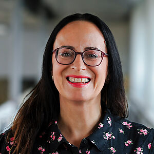 Portraitfoto von Iris Wemme-Baranowski. Sie lächelt mit offenem Mund. Ihr dunkles Haar fällt ihr über die Schultern. Sie trägt eine dunkelrote Brille und pinken Lippenstift. Sie ist in eine schwarze Bluse mit Blumenaufdruck gekleidet. Der Hintergrund ist verschwommen.