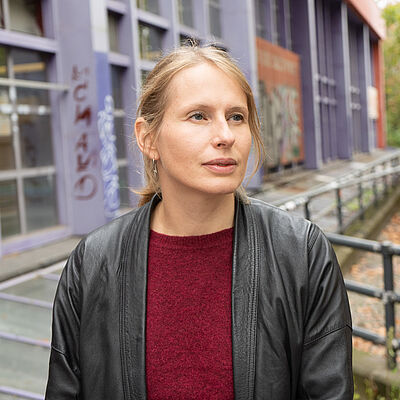 Portraitfoto von Mari-Liis Tigasson.  Sie sitzt draußen vor einem Gebäude. Sie trägt einen roten Pullover und darüber eine schwarze Lederjacke. Ihre  blonden Haare sind zu einem Zopf nach hinten gebunden.