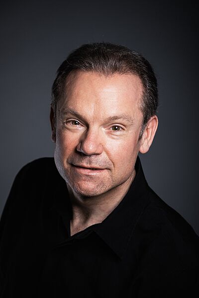 Portraitfoto von James Bobby. Er schaut von unten nach oben direkt in die Kamera. Er trägt sein Haar kurzgeschoren und ein schwarzes Hemd. Der Hintergrund ist grau.