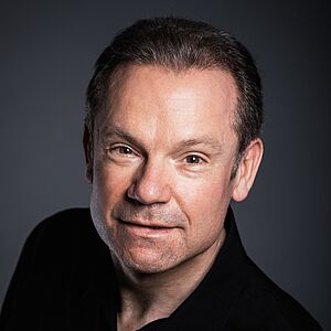 Portraitfoto von James Bobby. Er schaut von unten nach oben direkt in die Kamera. Er trägt sein Haar kurzgeschoren und ein schwarzes Hemd. Der Hintergrund ist grau. 