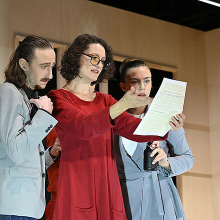 Drei Personen – die Frau im roten Kleid und mit Brille – starren auf einen Zettel. Sie halten Bier in der Hand und wirken erschrocken