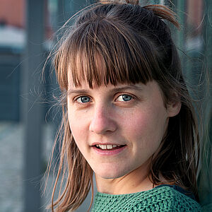 Portraitfoto von Meike Lehmann. Sie steht seitlich und und hat den Kopf zur Kamera gewandt. Ihr Pony fällt ihr über die Stirn. Sie hat dunkelblondes schulterlanges Haar. Sie trägt einen grünen gestrickten Pullover.