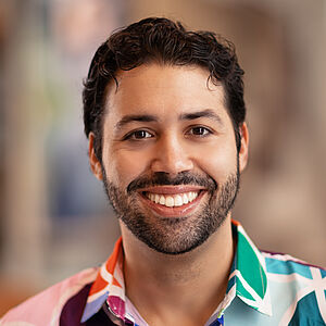Portraitfoto von Mario Orlando El Fakih Hernández. Er hat schwarzes Haar, einen Dreitagebart und trägt ein bunt gemustertes Hemd. Er lächelt mit geöffnetem Mund. 