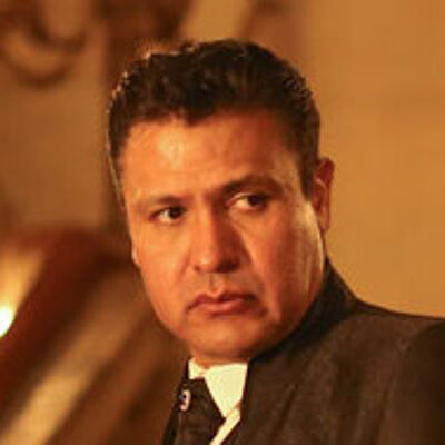 Portraitfoto von Juan Orozco. Sein Haar ist zurückgegelt. Er blickt zur Seite. Er trägt einen schwarzen Anzug mit schwarzer Krawatte und weißem Hemd. Im Hintergrund ist verschwommen ein Treppenhaus mit gewundenem Geländer zu erahnen.