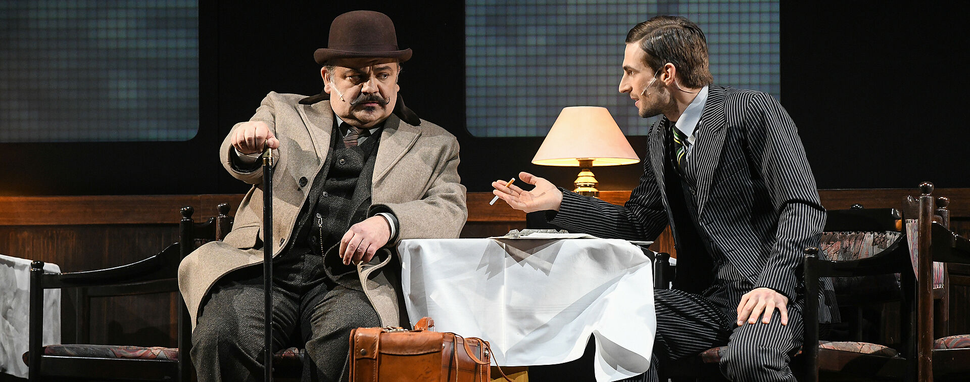 Hercule Poirot und Samuel Ratchett sitzen sich an einem Tisch gegenüber. Poirot schaut skeptisch. Seine rechte Hand lehnt auf einem Gehstock. Er trögt einen beigen Mantel, einen Tweedanzug, eine braune Melone auf dem Kopf und einen Schnauzer im Gesicht. Sein linker Arm lehnt auf der weißen Tischdecke. Ratchett trägt einen dunkelgrauen Nadelstreifenanzug, dazu schwarz-weiße Budapester und zurückgegeltes Haar. Sein rechter Arm lehnt auf dem Tisch, in der Hand eine Zigarette.