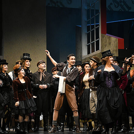 Zu erkennen ist eine große Personengruppe. Im Mittelpunkt steht der Protagonist, der ein helles Küchentuch über dem rechten Arm hält.