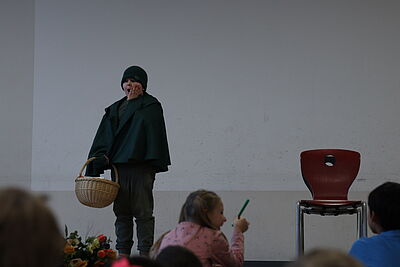 Ein Junge mit einem grünen Umhang und einem geflochtenen Körbchen steht mit geöffnetem Mund und davor gehaltener Hand vor dem Publikum.