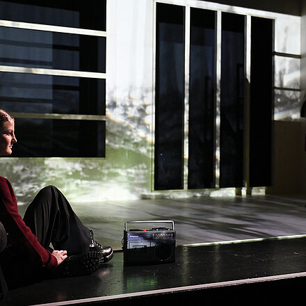 Links sitzt ein Mädchen mit einem Radio. Im Hintergrund ist das Bühnenbild mit einer Landschaft zu erkennen. Hinten steht die Frau im roten Kleid.